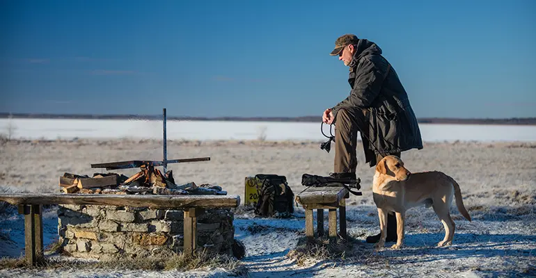 En person står med foten på en bänk. En hund står nedanför. Det är snö och vitt på marken.