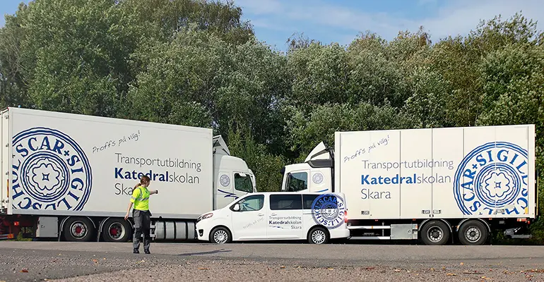En tjej som pekar bakåt mot två stora lastbilar och en minibuss, som alla är dekorerade med Katedralskolans sigill och texten "Transportutbildning Katedralskolan Skara".