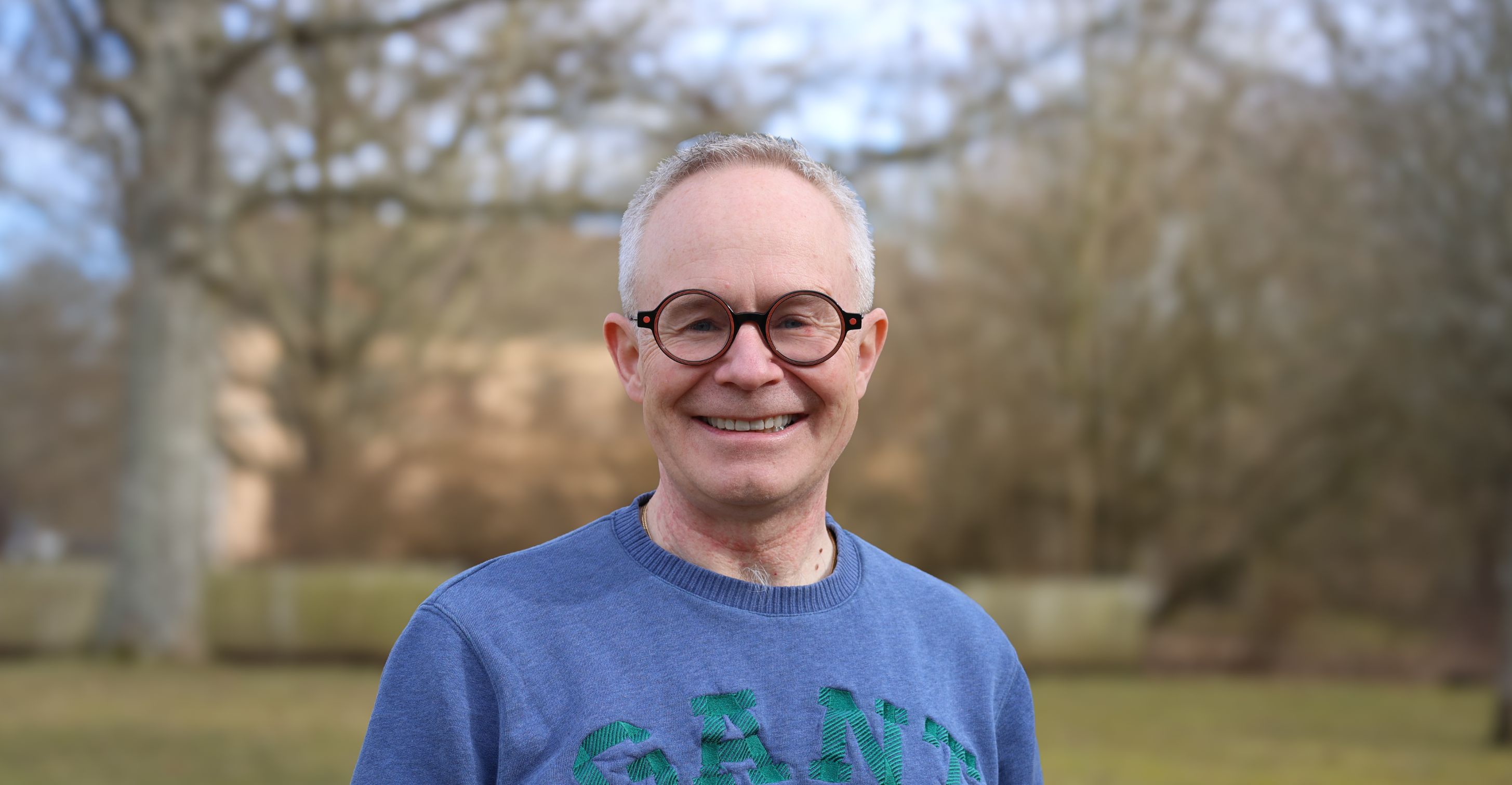 Porträtt av en leende man med runda glasögon och blå tröja i en utomhusmiljö.