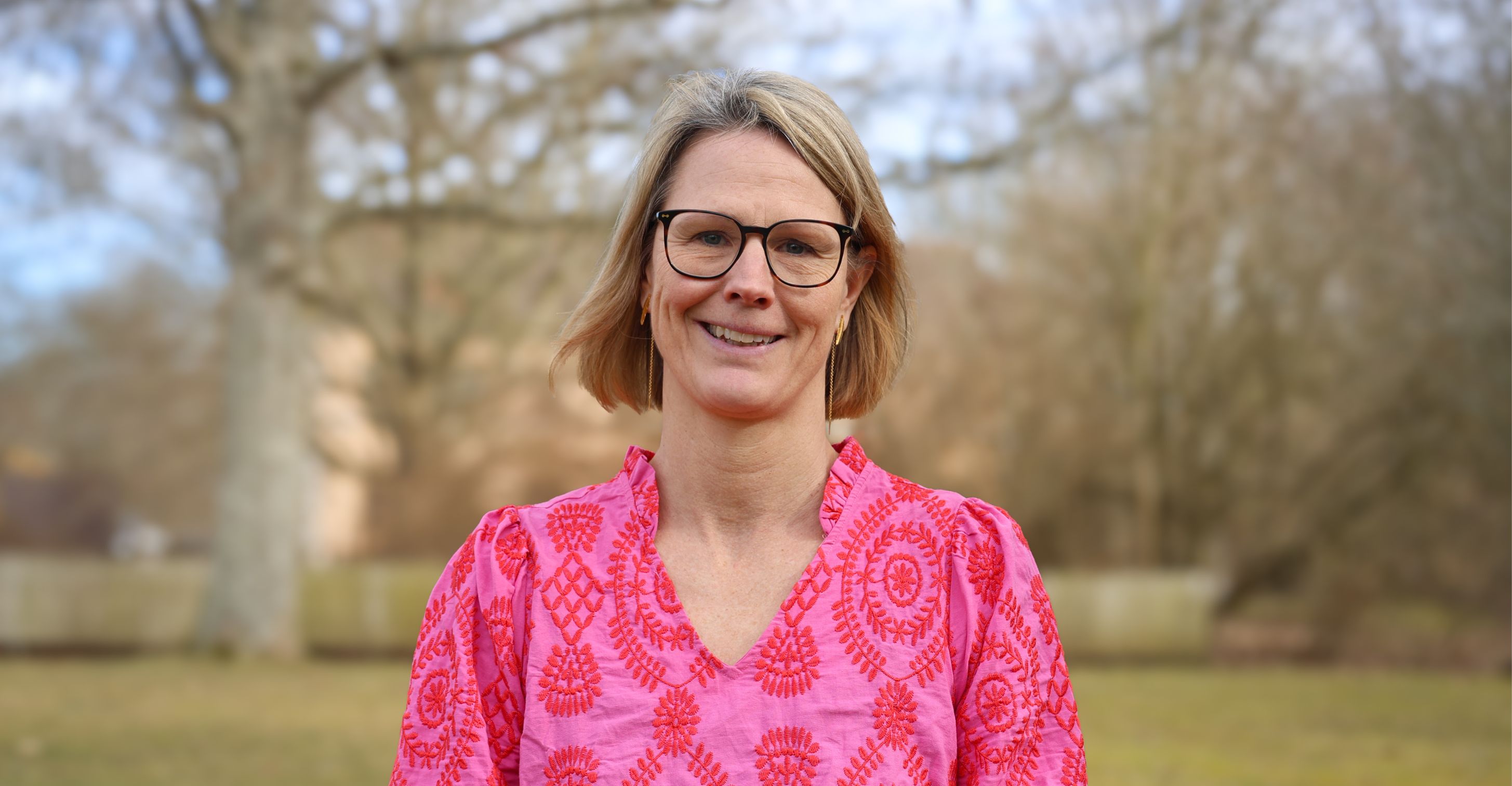 Porträtt av en leende kvinna i rosa blus utomhus under en molnig dag.