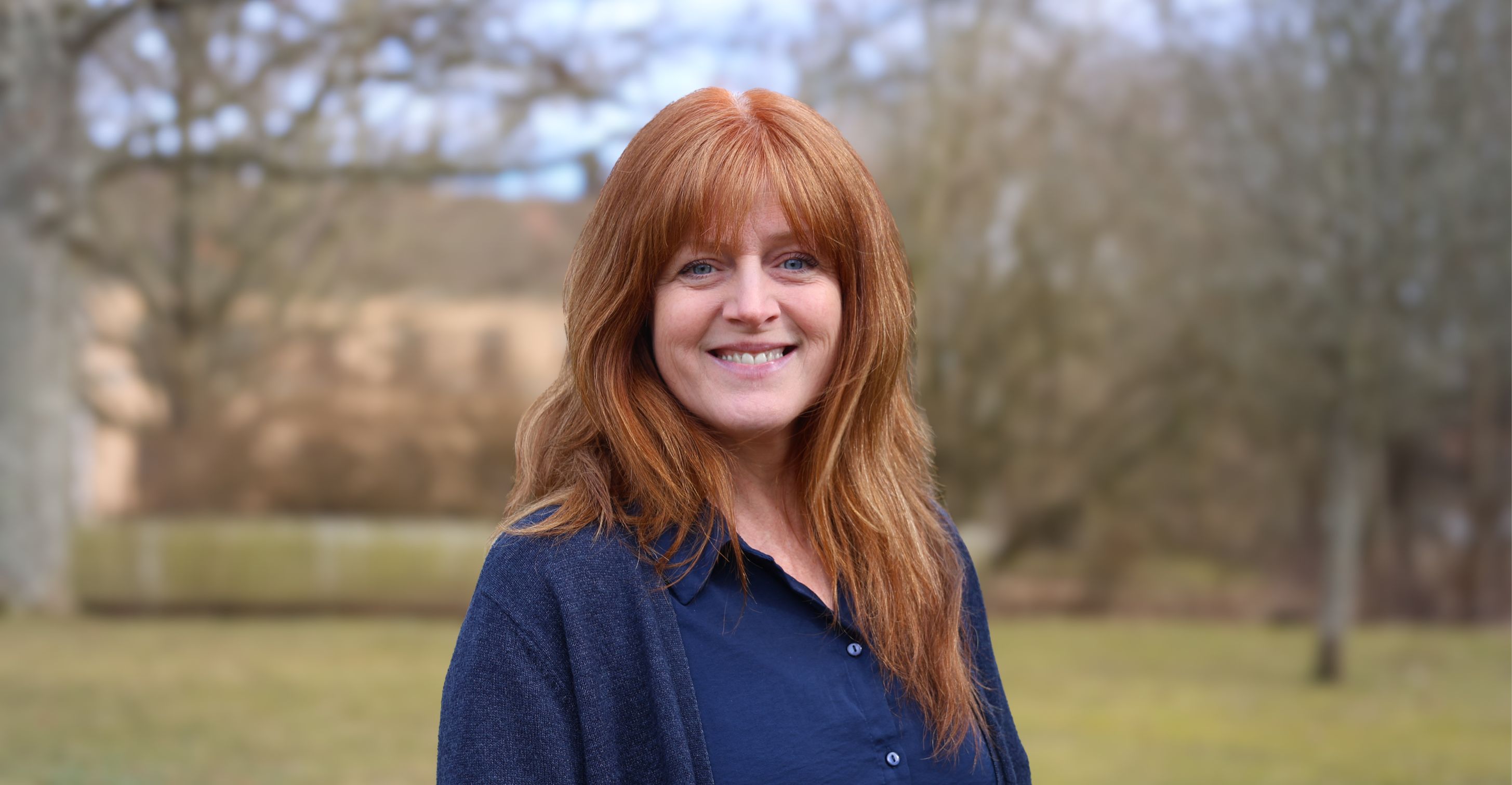 Porträtt av en leende rödhårig kvinna i mörkblå kläder utomhus.
