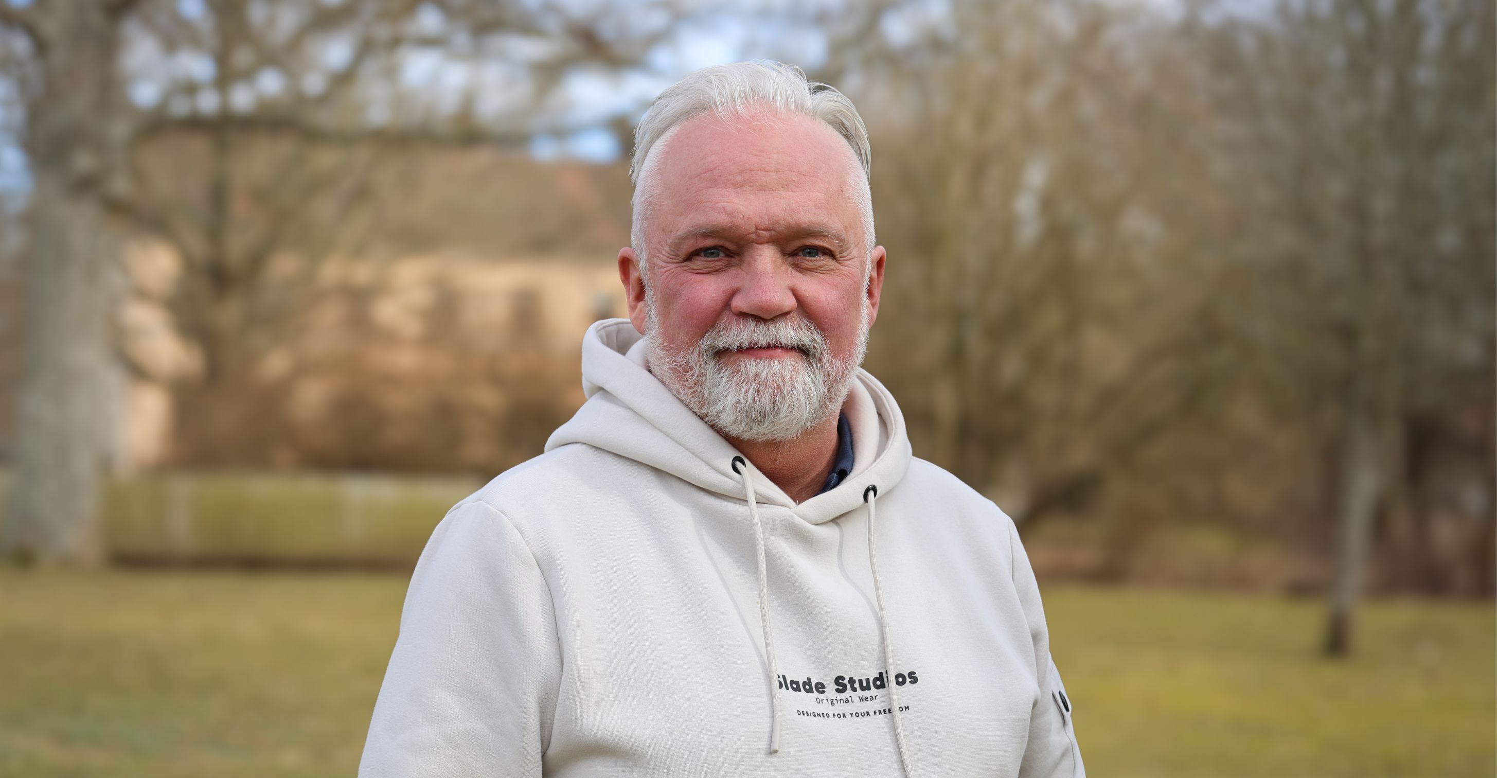 Porträtt av en man med grått skägg klädd i en ljus hoodie, fotograferad utomhus.