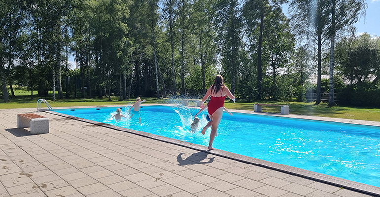 Flera barn badar i en bassäng och ett barn är på väg att hoppa i. Det är soligt och man ser träd i bakgrunden.