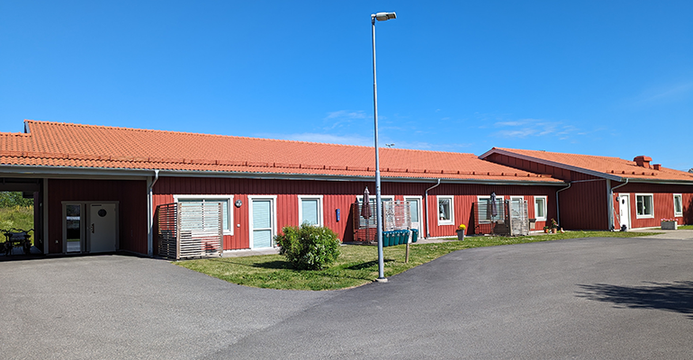Rött enplans hus med flera lägenheter. Uteplats och liten gräsmatta framför varje lägenhet, bild.